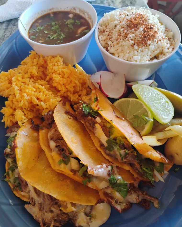 Birria platter — orange-stained tacos with a cup of consommé and rice on a blue plate