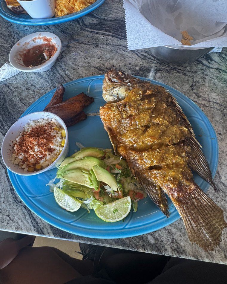 Whole fried mojarra — a golden, crispy whole fish with sliced avocado and lime on a blue plate