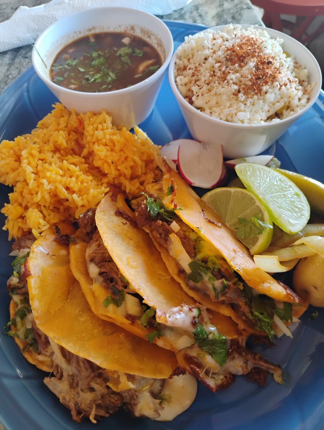 Birria tacos stained deep orange with a cup of rich consommé and rice on a blue plate