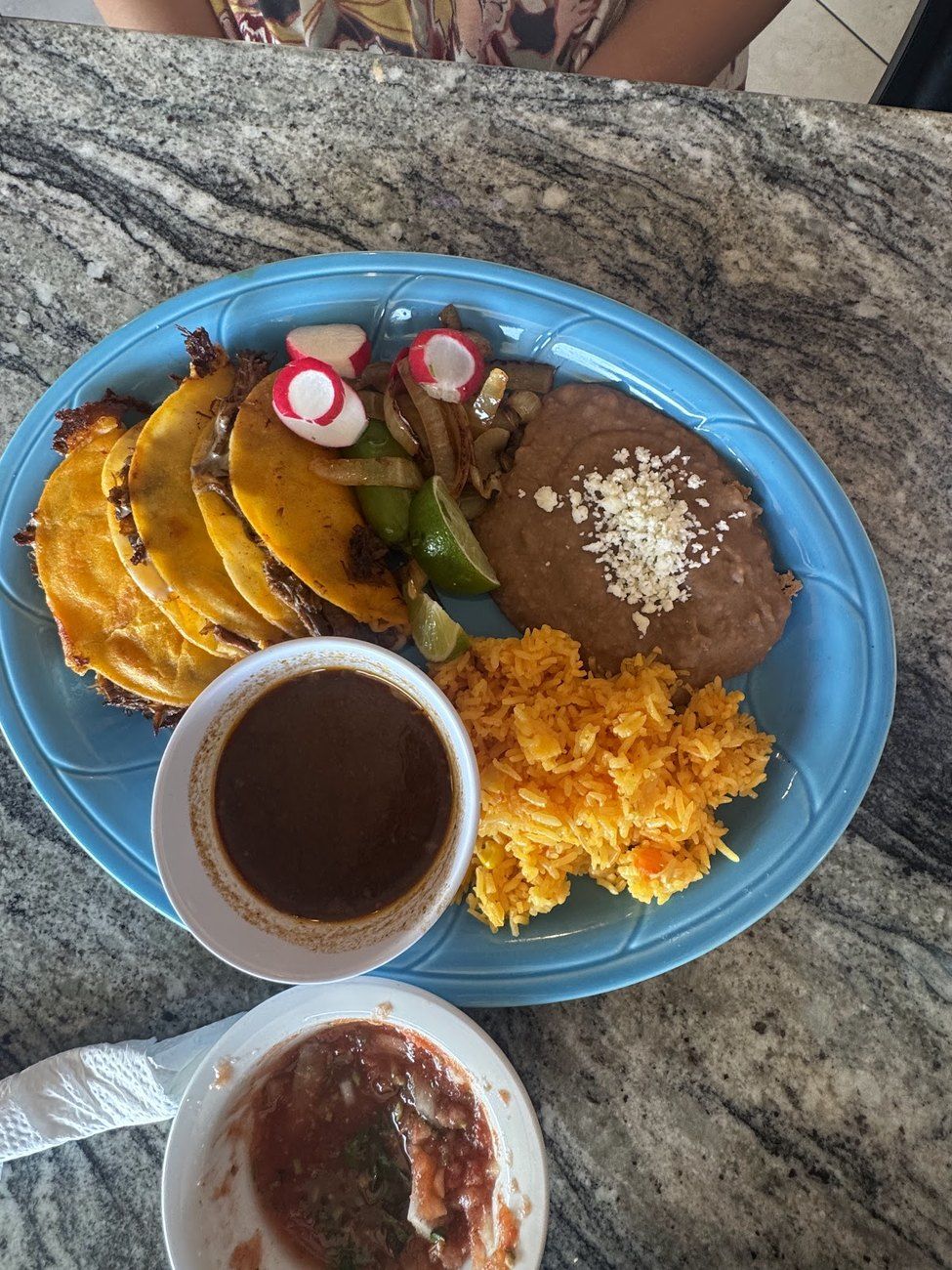 Birria tacos with a cup of consommé on a blue plate, set outdoors